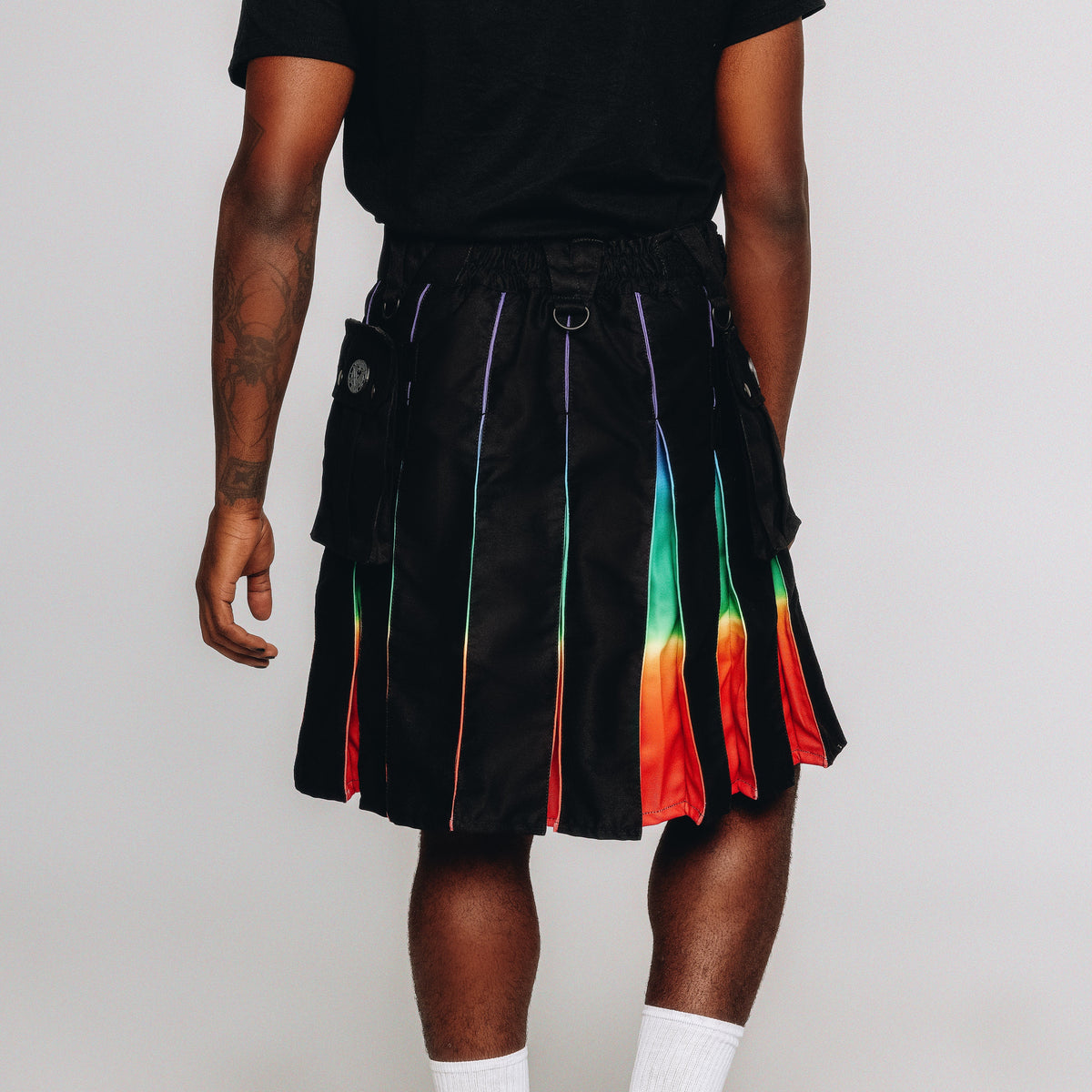 A model from the back wearing a black pleated skirt with vertical rainbow-colored stripes, styled with a black t-shirt, white socks, and black shoes against a light gray studio background.