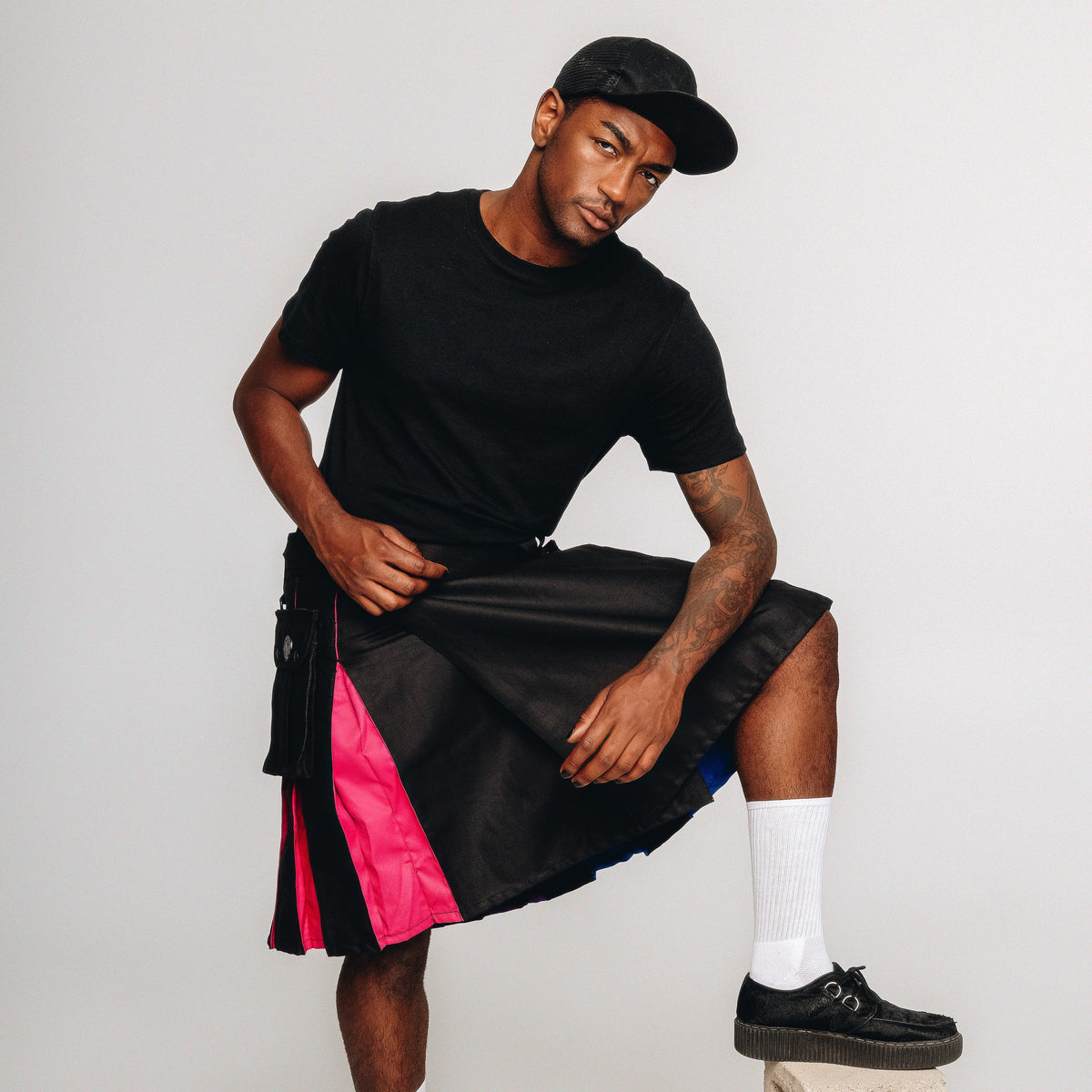 A model wearing a black t-shirt, a black cap, and a black kilt with vibrant rainbow side panels, paired with white socks and black shoes, posing against a light gray studio background.