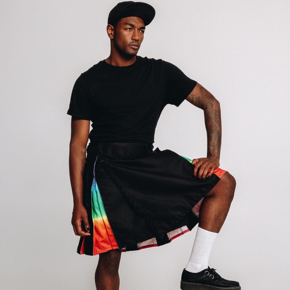 A man models a black t-shirt and black kilt with a vibrant rainbow stripe down the side, paired with white socks and black shoes, posing on a concrete block against a light gray background.
