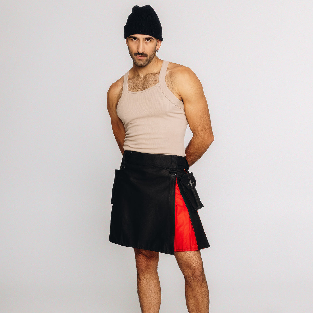 A man modeling a black wrap-style kilt with a bright red side panel, paired with a beige tank top and black beanie, against a plain light gray studio background.