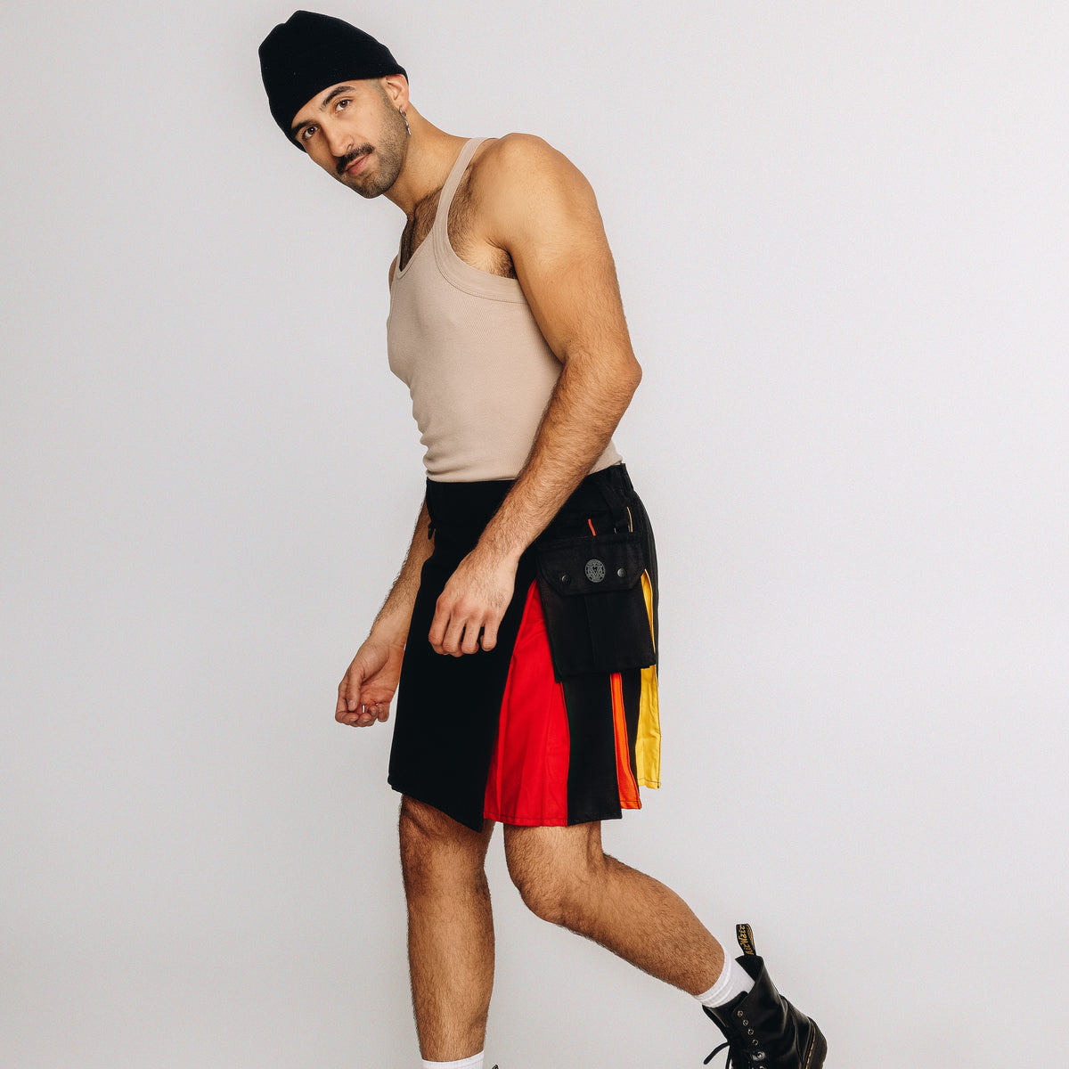 A man wearing a beige tank top, black beanie, and a black kilt with red, yellow, and green paneling walks against a light gray studio background.