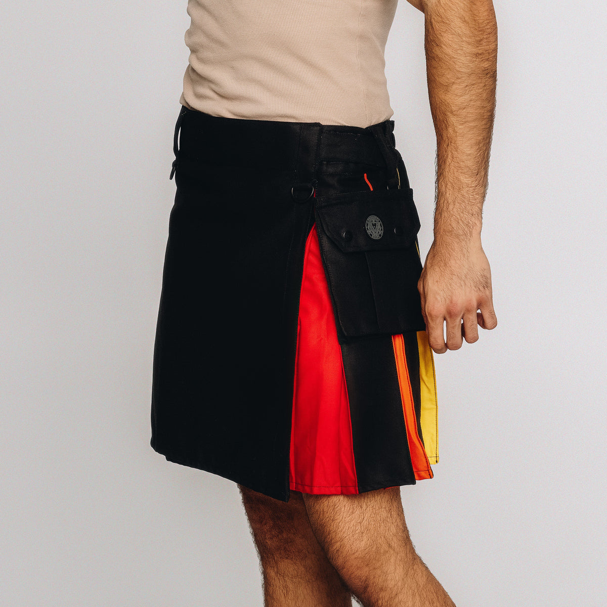 A man wearing a black leather kilt with vertical panels of red, orange, and yellow, paired with a beige t-shirt, shown from the chest down against a light gray background.