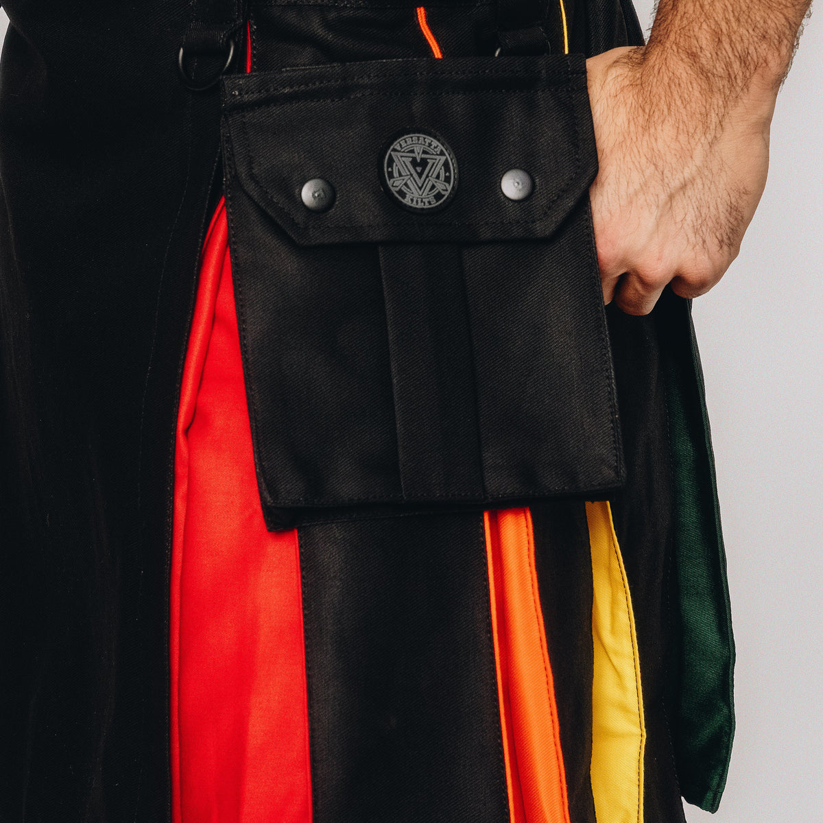 Close-up of a black fabric pouch with silver snap buttons and a circular metal emblem, worn over a garment with bold red, orange, yellow, and green vertical panels. A person's hand is visible holding the pouch.