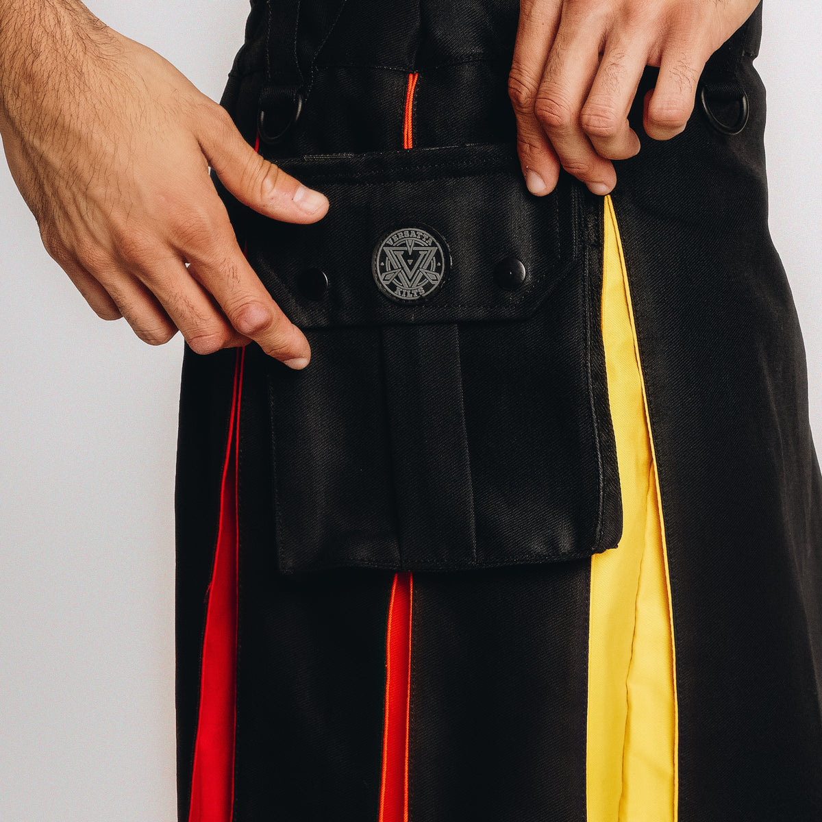 Close-up of a person wearing black pleated kilt with bright red and yellow vertical stripe accents, adjusting a front flap pocket with a circular metal emblem.