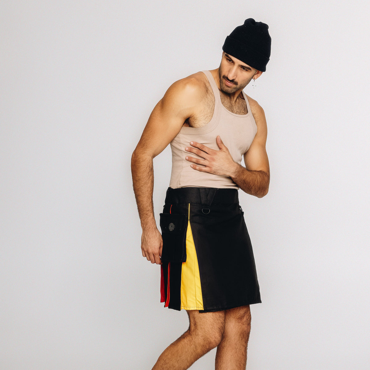 A man modeling a black leather kilt with yellow and red side panels, paired with a beige tank top and black beanie, against a plain light gray studio background.