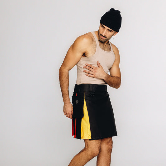 A man modeling a black leather kilt with yellow and red side panels, paired with a beige tank top and black beanie, against a plain light gray studio background.
