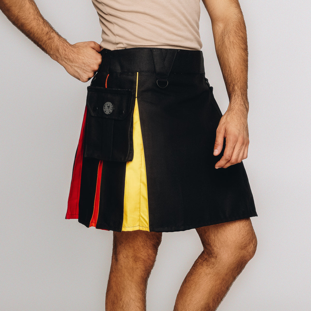 A man wearing a black kilt with vertical panels of red, yellow, and black, featuring a black utility pouch on the front, photographed against a light gray studio background.