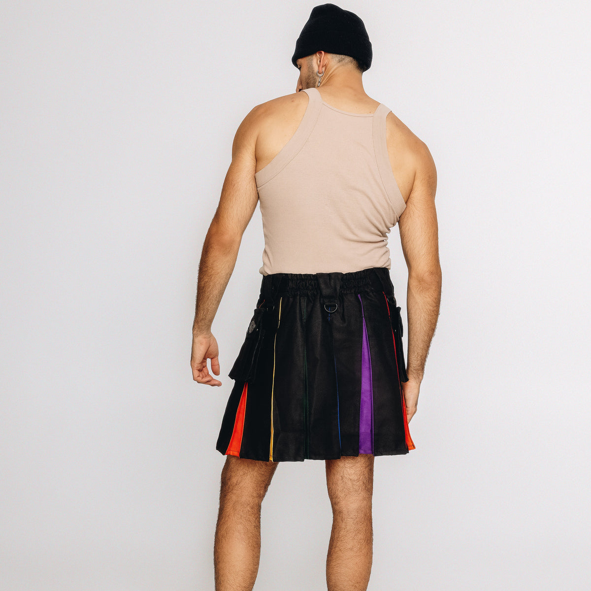 A man shown from behind wearing a beige ribbed tank top, a black beanie, and a black pleated kilt with vertical rainbow-colored stripe panels on the sides, set against a plain light gray studio background.
