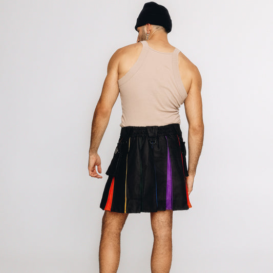 A man shown from behind wearing a beige ribbed tank top, a black beanie, and a black pleated kilt with vertical rainbow-colored stripe panels on the sides, set against a plain light gray studio background.