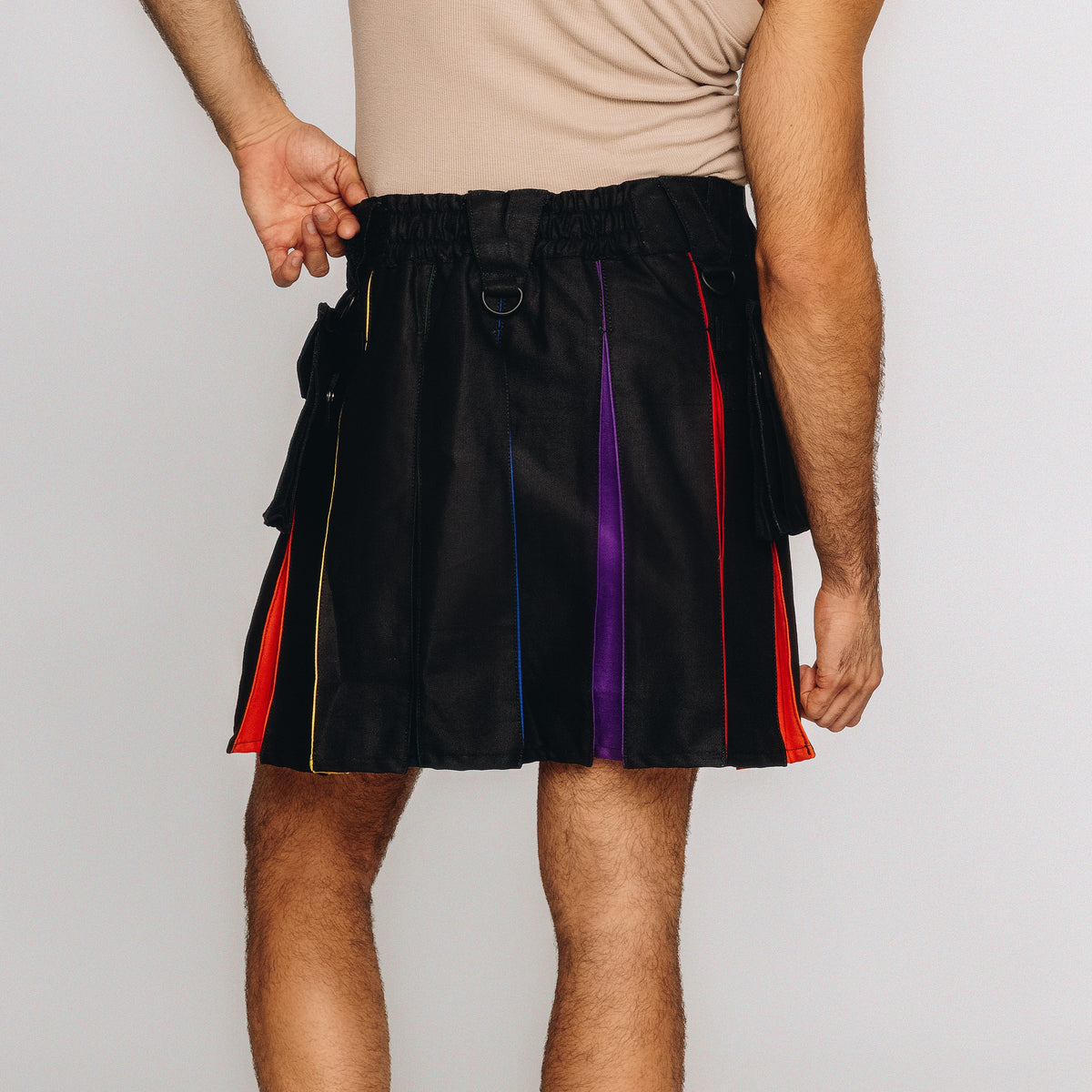 A person wearing a black pleated kilt with vertical rainbow-colored stripes in red, orange, yellow, blue, and purple, paired with a beige tank top against a light gray studio background.