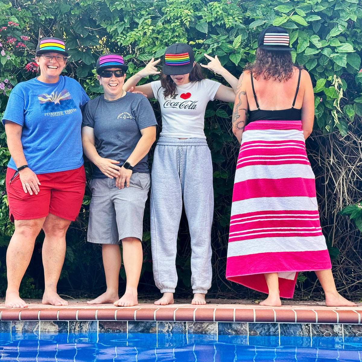 Four people standing on the edge of a swimming pool in front of a lush green hedge, wearing colorful striped hats and casual summer clothing.