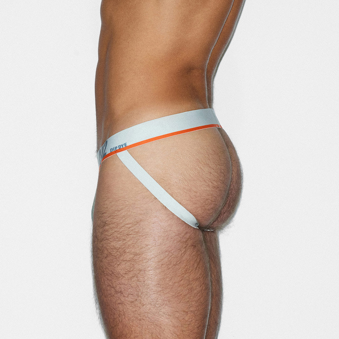 A close-up side view of a male model wearing a white jock-style brief with a thin orange stripe and a small blue C-IN2 brand logo on the waistband, against a light gray studio background.