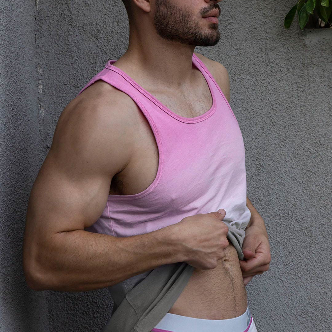 Man wearing a pink tank top and gray shorts against a gray wall with greenery.