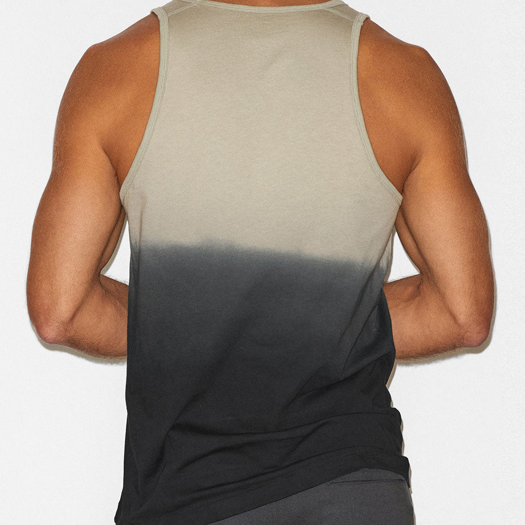 A man shown from the back wearing a sleeveless tank top with a vertical ombre gradient transitioning from light beige at the top to dark charcoal gray at the bottom, paired with dark gray shorts against a plain white background.