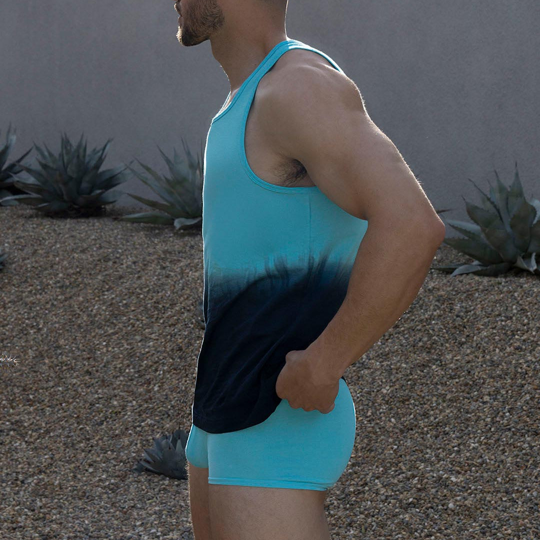 Man wearing a gradient blue tank top and shorts standing outdoors with plants in the background