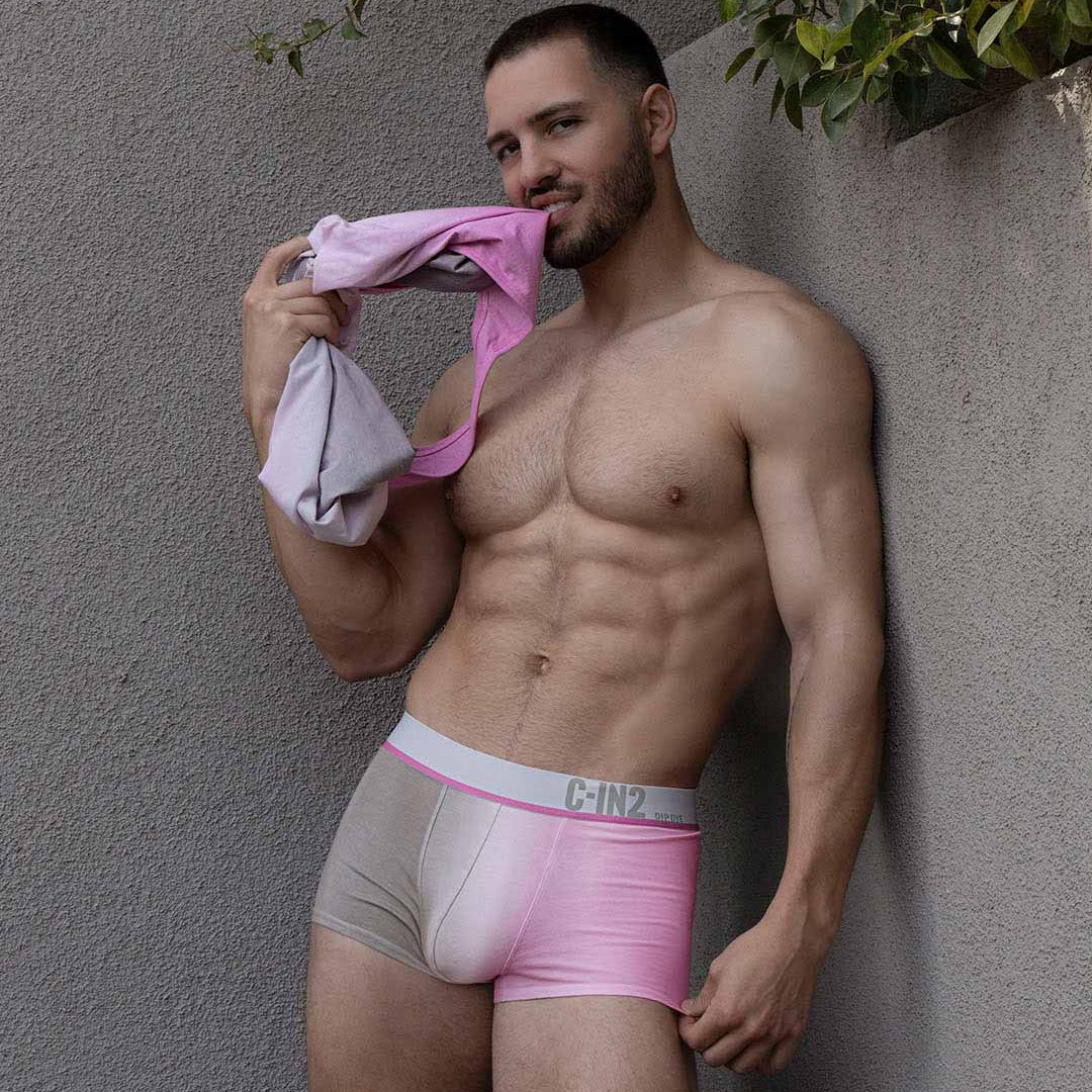 Man wearing pink underwear with C-IN2 branding with a gray waistband, holding a pink shirt against a gray wall.