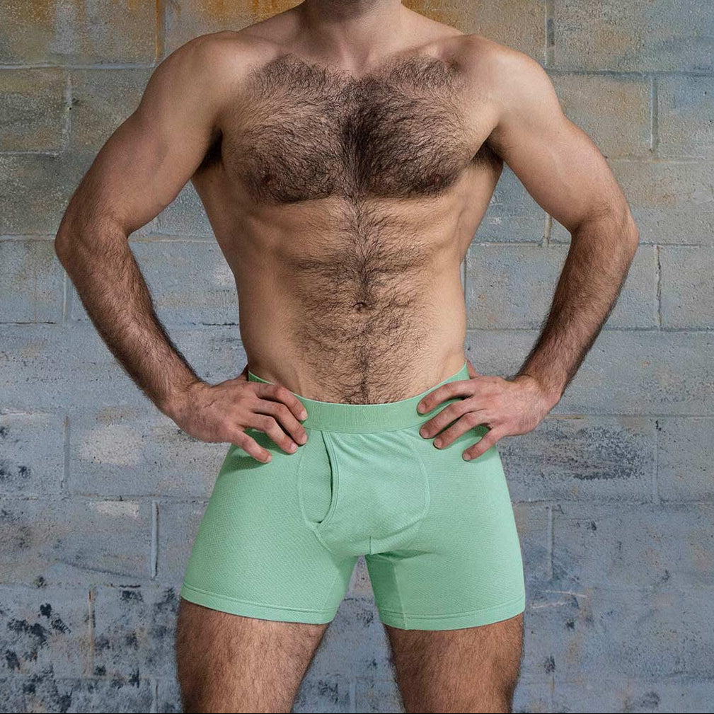 Man wearing light green boxer briefs against a textured wall.