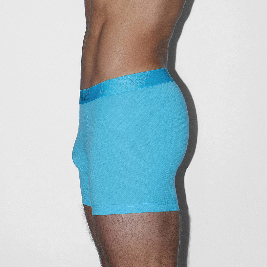 A side profile view of a man wearing bright blue boxer briefs against a plain light gray background.