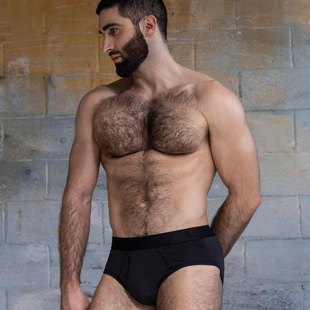 Man wearing black underwear against a textured wall.