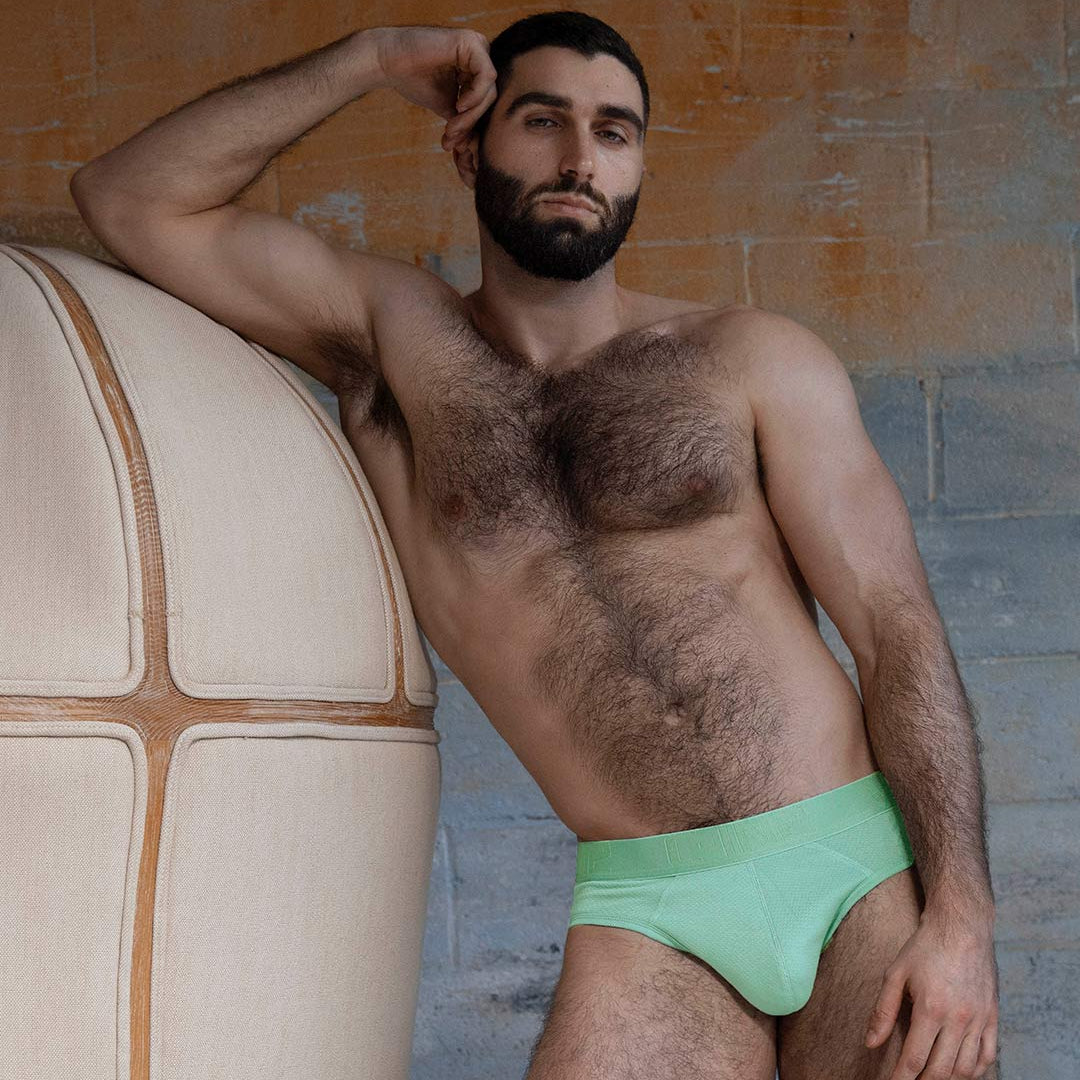 Man wearing green underwear leaning against a textured wall.