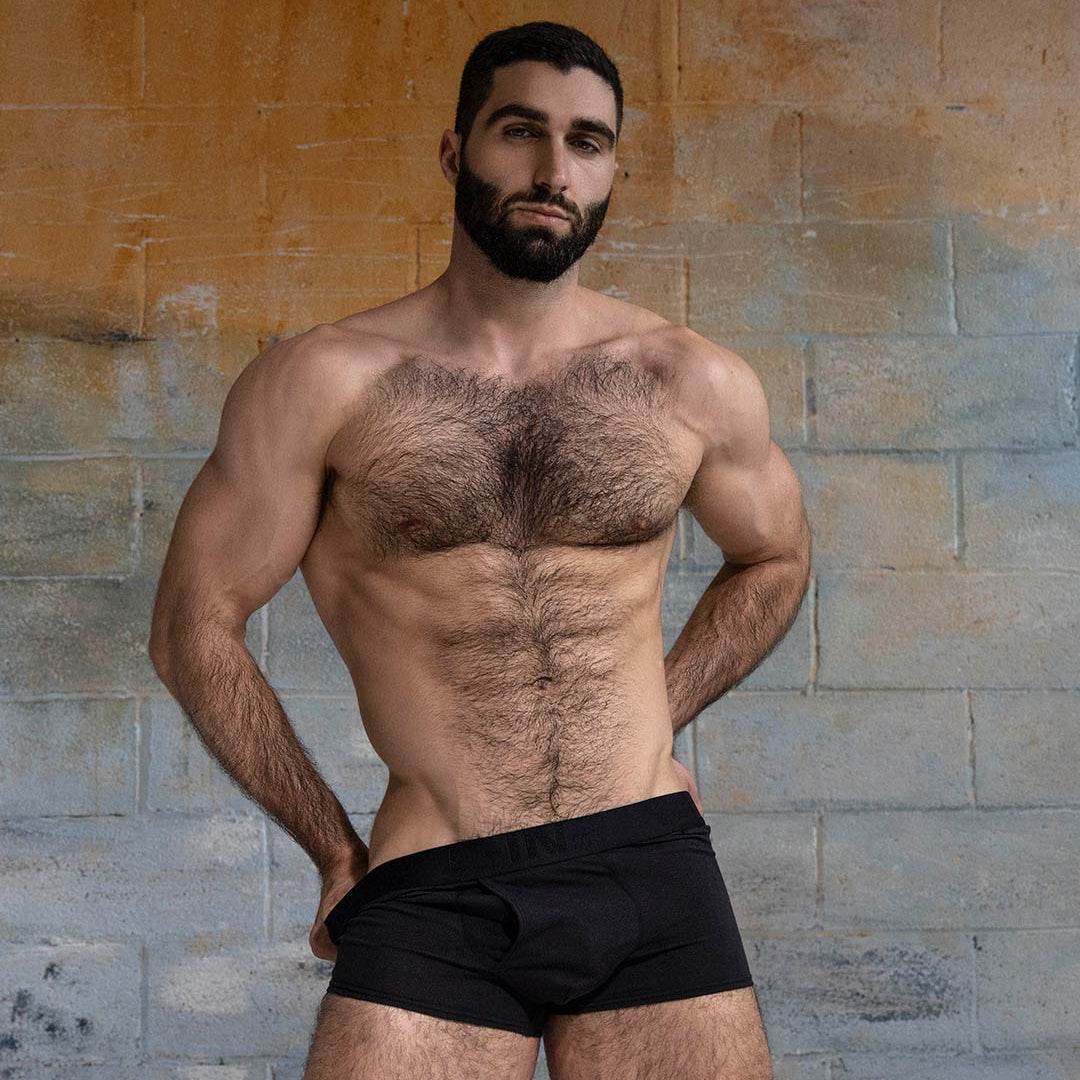 Man wearing black underwear against a textured wall.