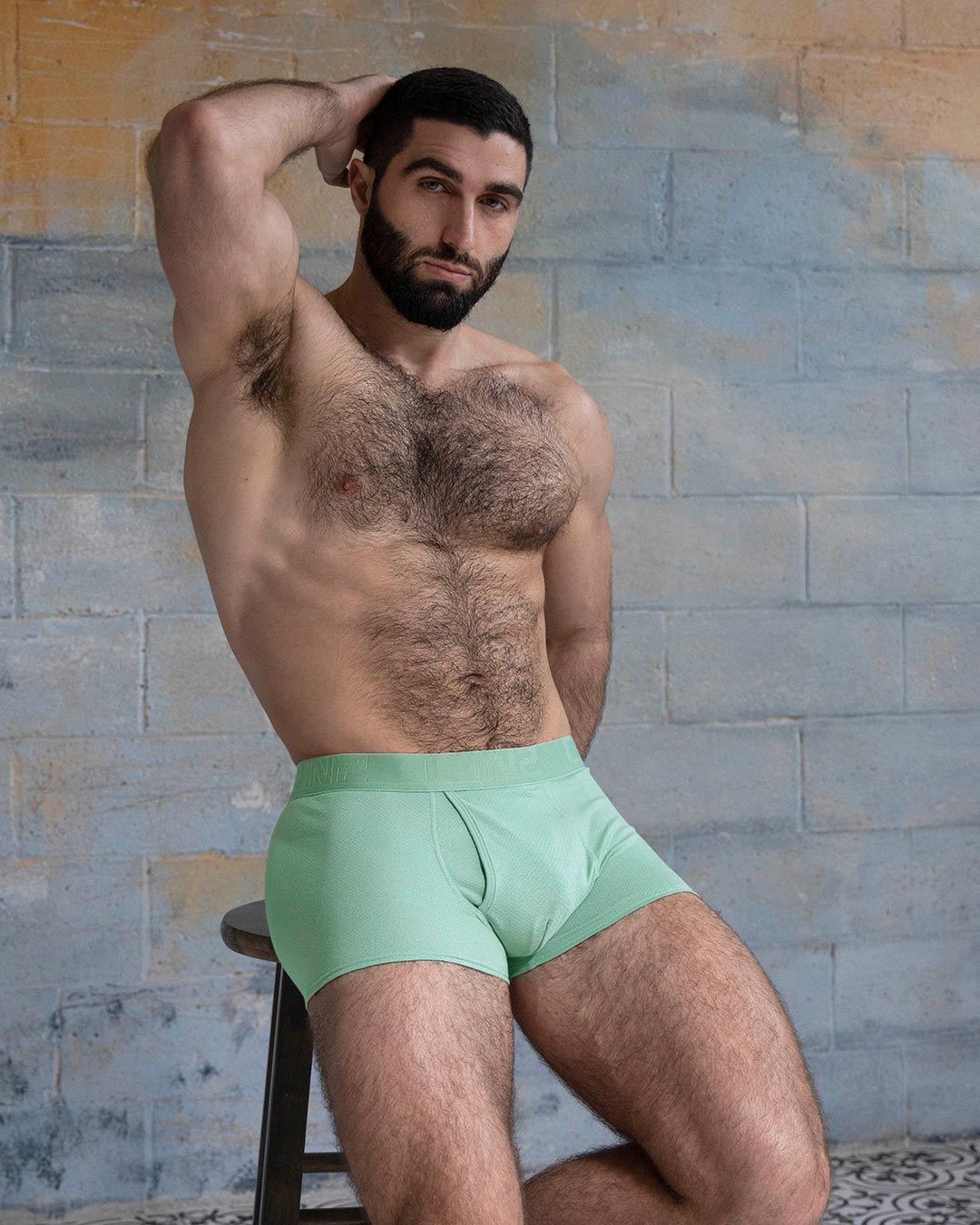 Man wearing light green boxer briefs against a textured wall.