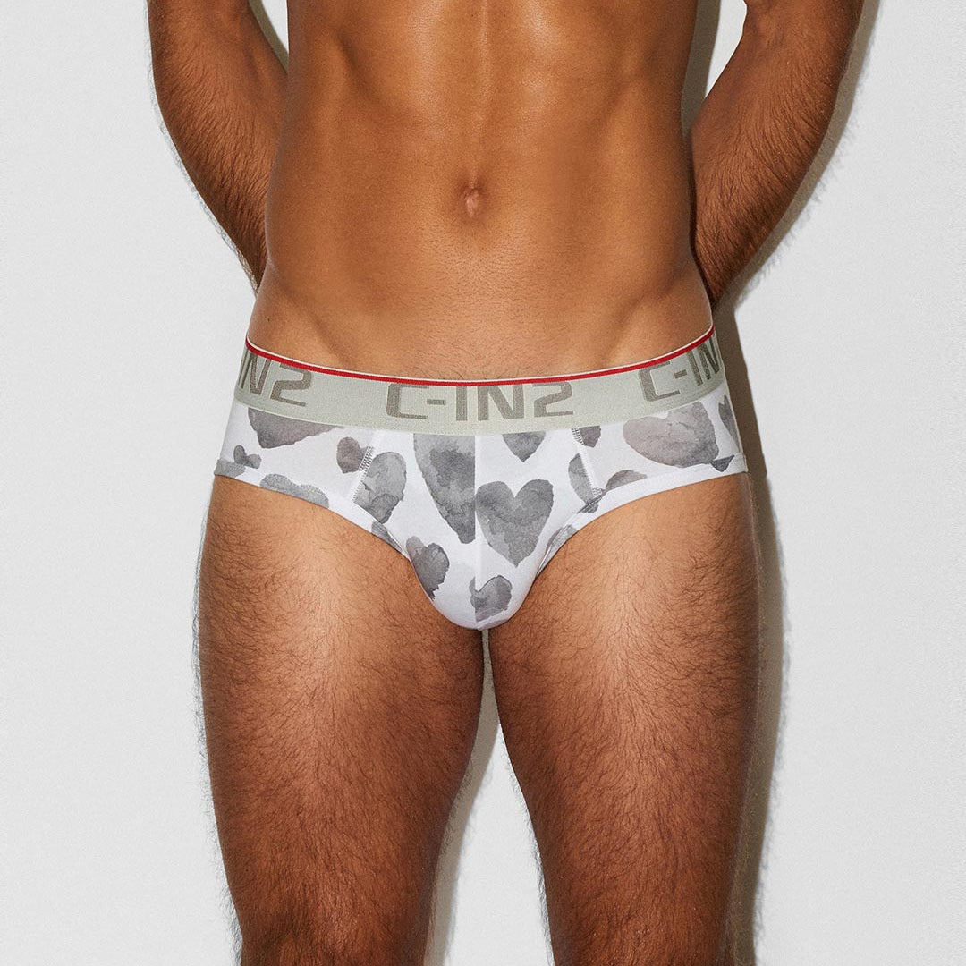 A close-up of a male model's lower torso wearing low-rise underwear. The briefs are white with a gray heart pattern and a gray waistband featuring the brand name 'C-IN2' in gray lettering with a red accent line.