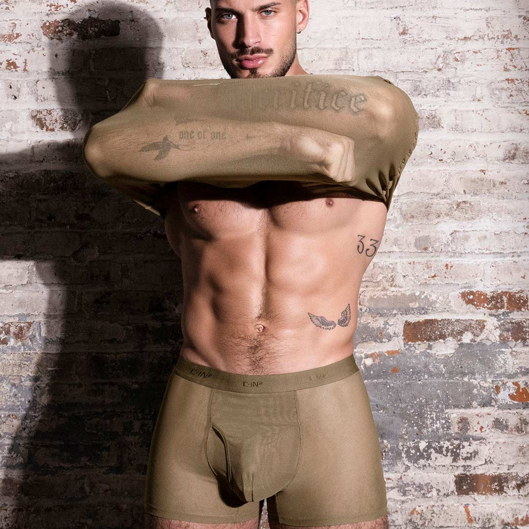 A male model wearing olive green underwear and a matching olive green shirt pulled up over his chest, standing against a distressed white brick wall.