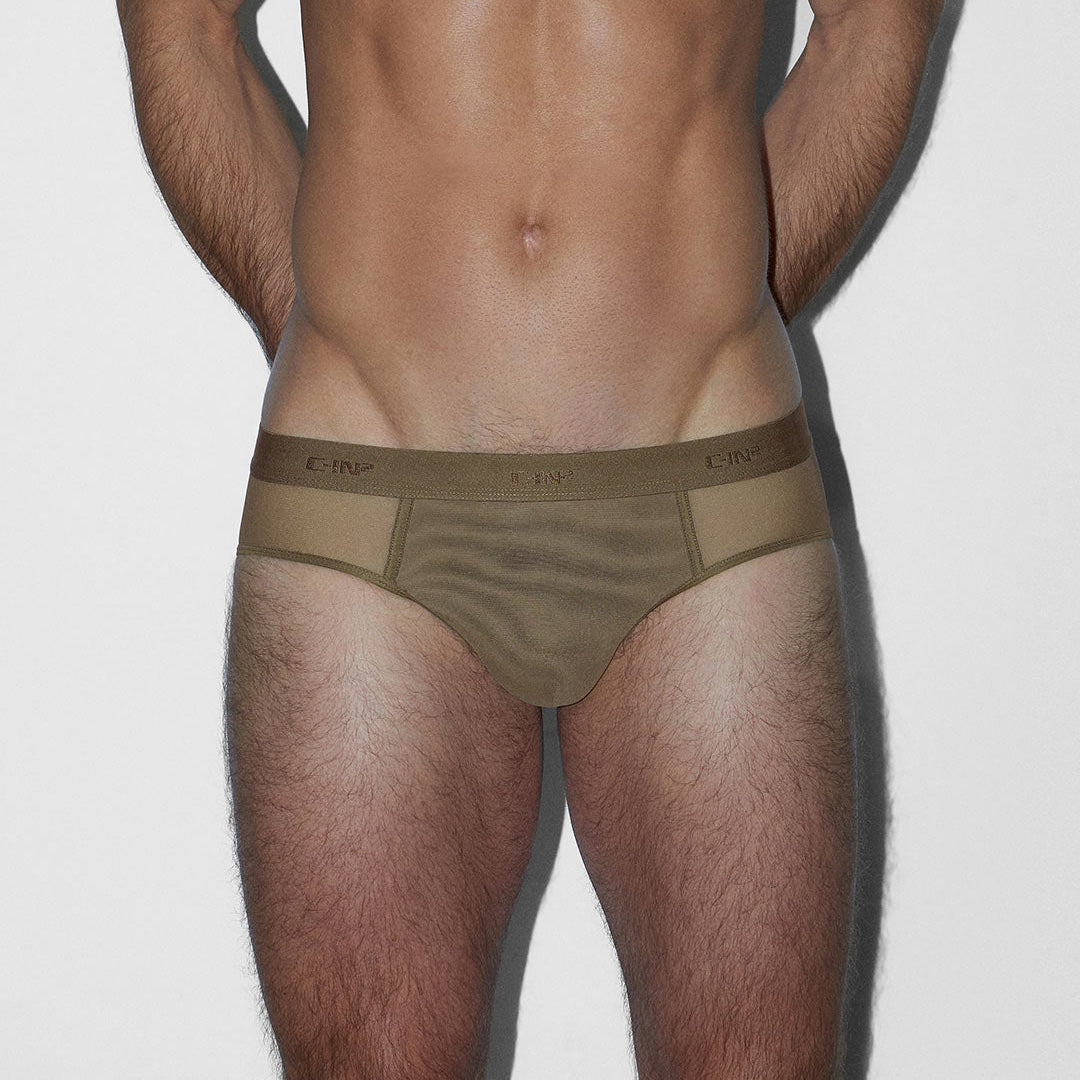 A man's lower torso and thighs wearing low-rise briefs in a muted taupe-brown color. The underwear features a wide waistband with 'C-IN2' branding repeated on it, and a sheer mesh front panel.