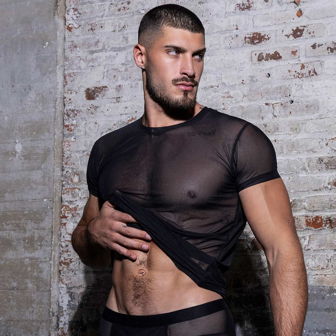 Man wearing a sheer black mesh top and underwear against a textured wall.