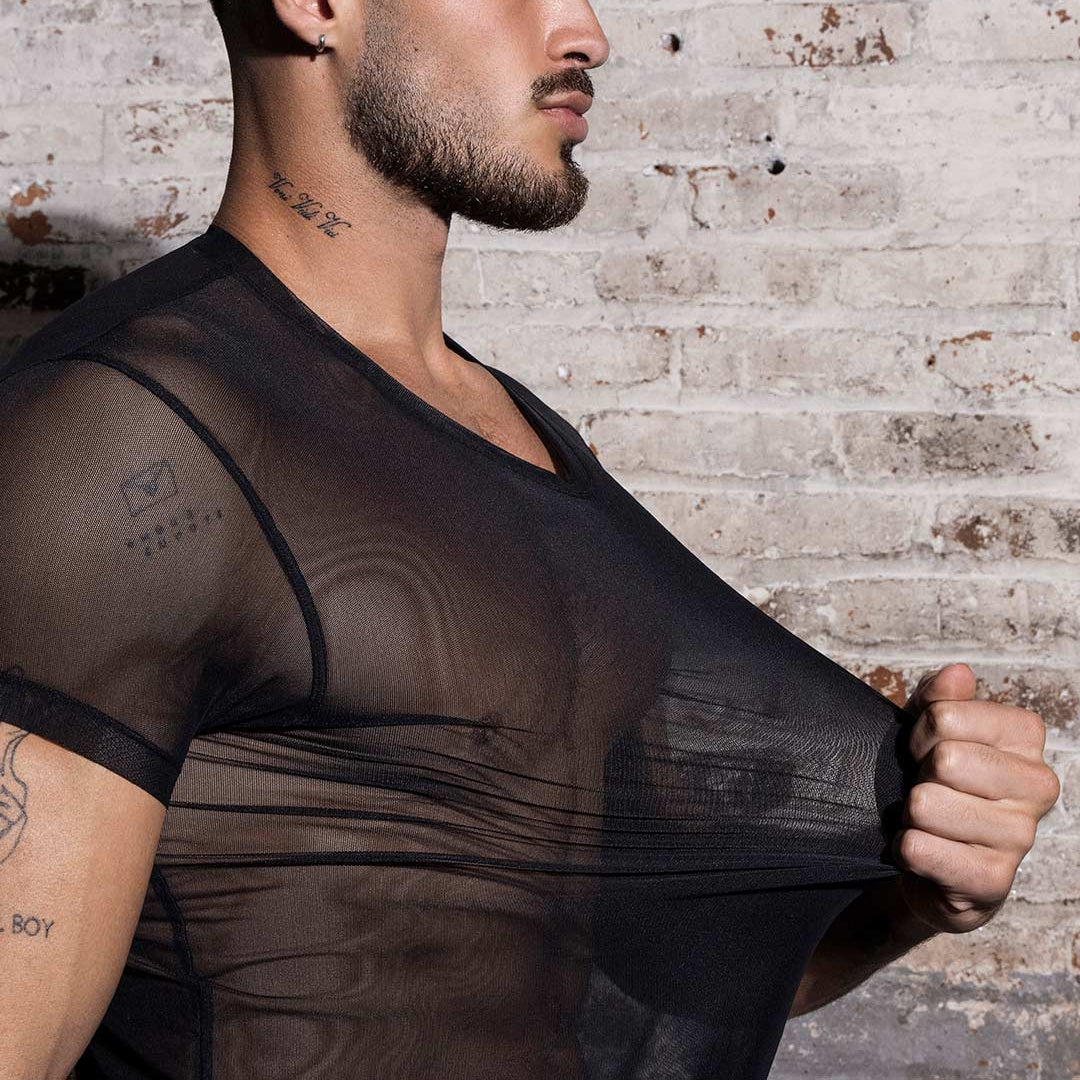 A man wearing a sheer black mesh crew-neck t-shirt, pulling the fabric outward to demonstrate its stretch, set against a rustic white brick wall background.
