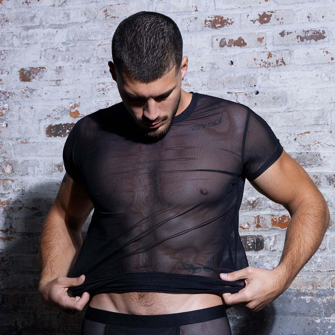 A man wearing a sheer black short-sleeve mesh t-shirt and matching black mesh briefs, posing against a rustic white brick wall.