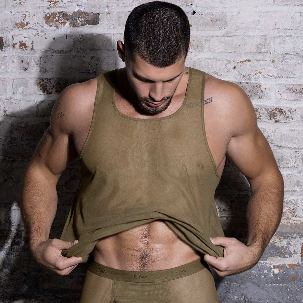 Man wearing a green sheer tank top set against a textured brick wall.
