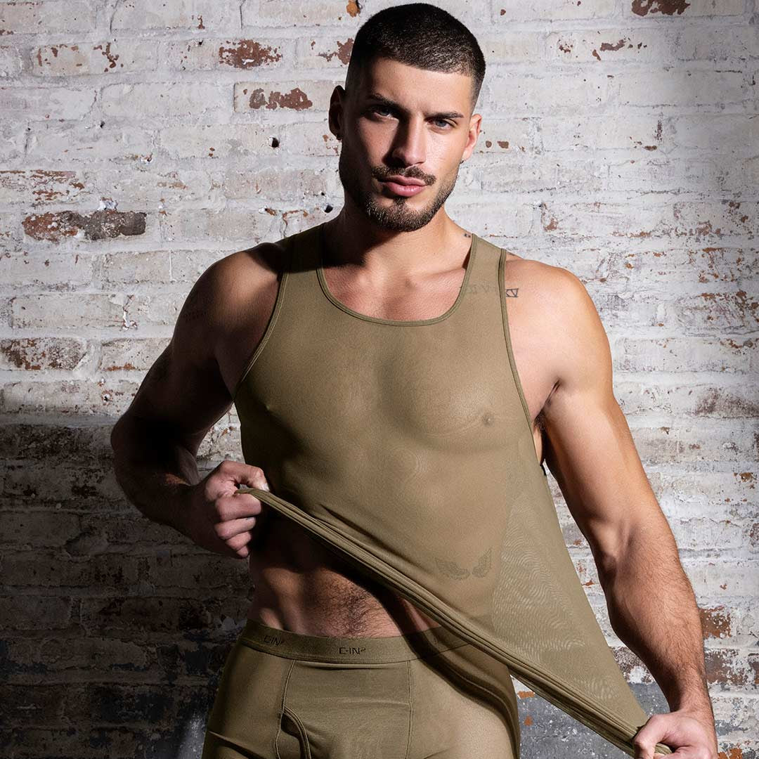 Man wearing a beige sheer tank top against a brick wall.