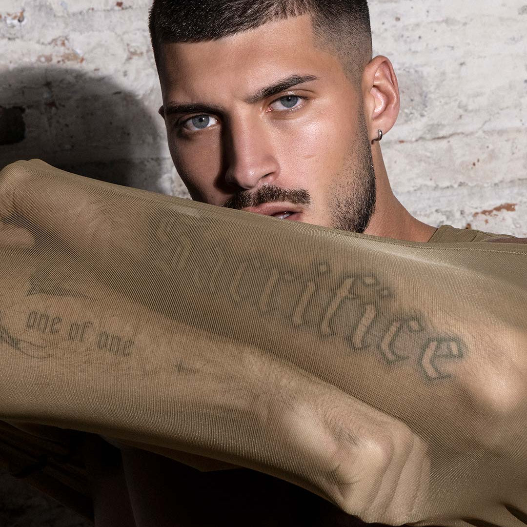 Man with tattoos holding a beige sheer shirt against a textured wall.