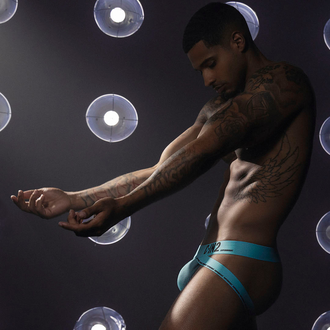 A tattooed male model wearing bright turquoise blue jock poses against a dark studio background with circular spotlight effects.