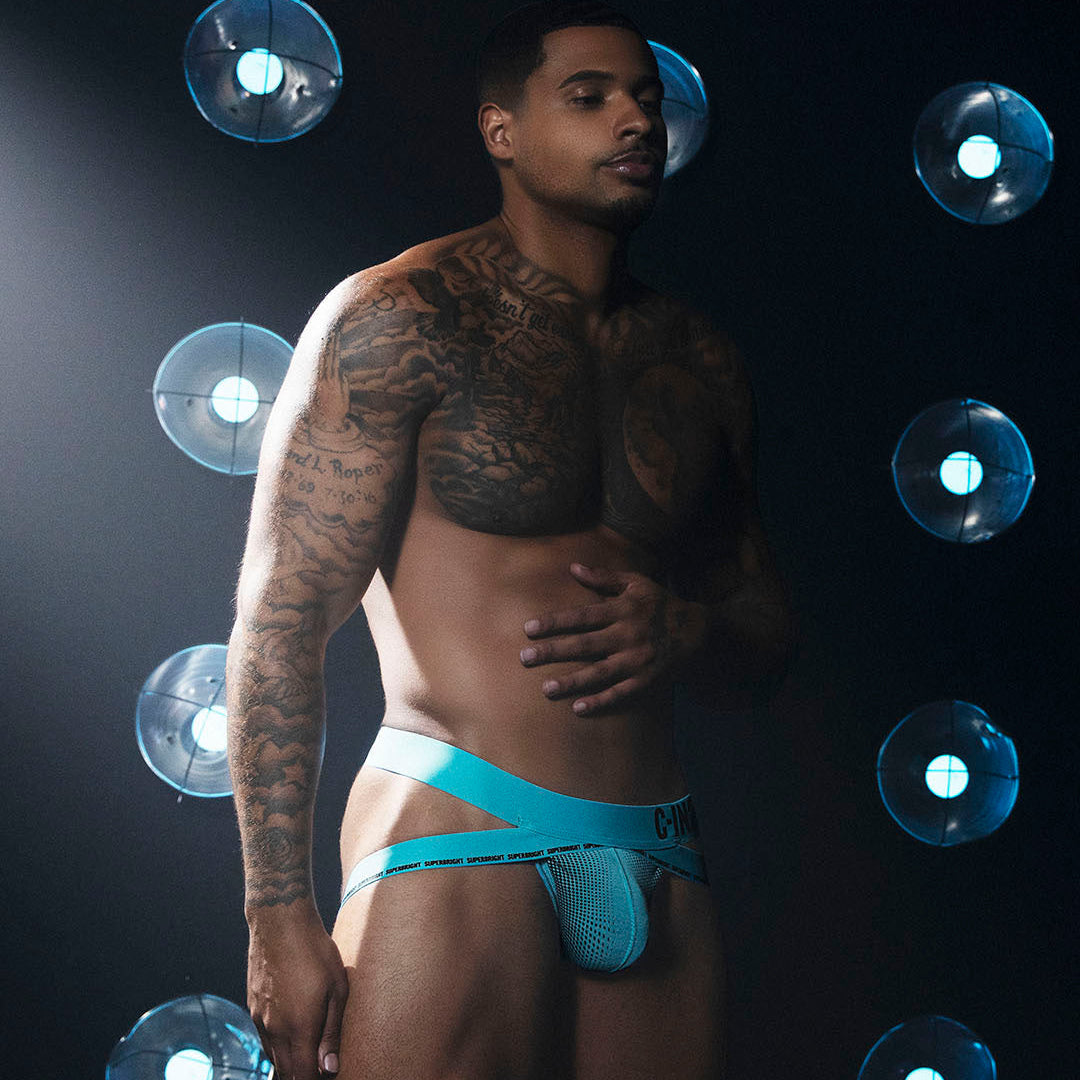 A tattooed male model wearing bright turquoise blue men's underwear, standing against a dark background with circular blue light fixtures.