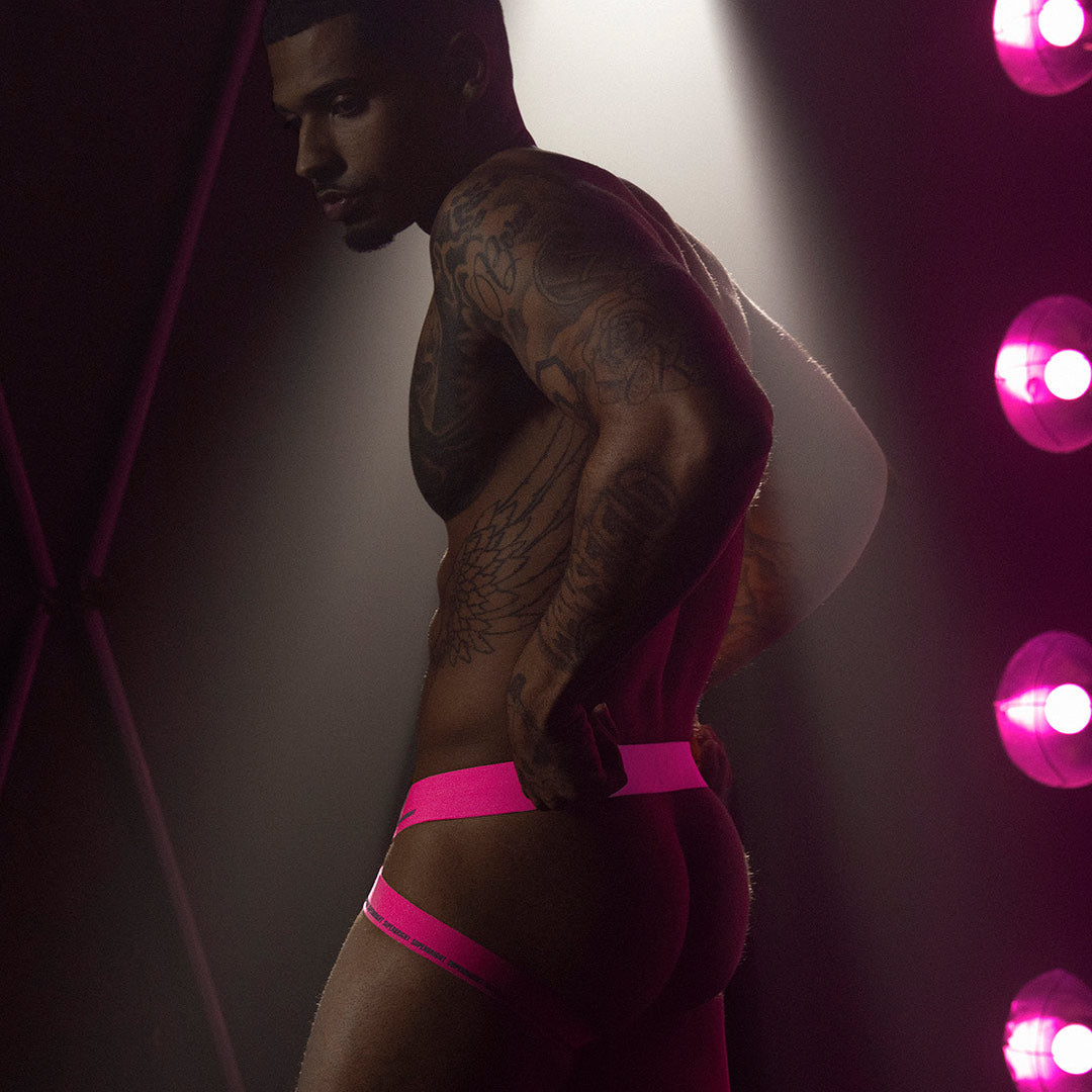 Man wearing pink jockstrap with a dark background and pink lights.