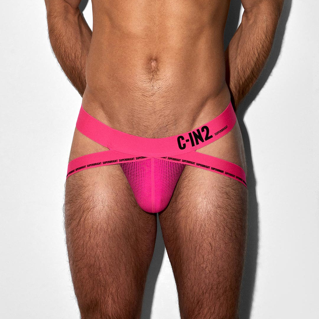 A close-up of a male model's lower torso wearing a bright hot pink jock-style brief with wide crisscross straps and black 'C-IN2' branding on the side, against a plain light gray background.