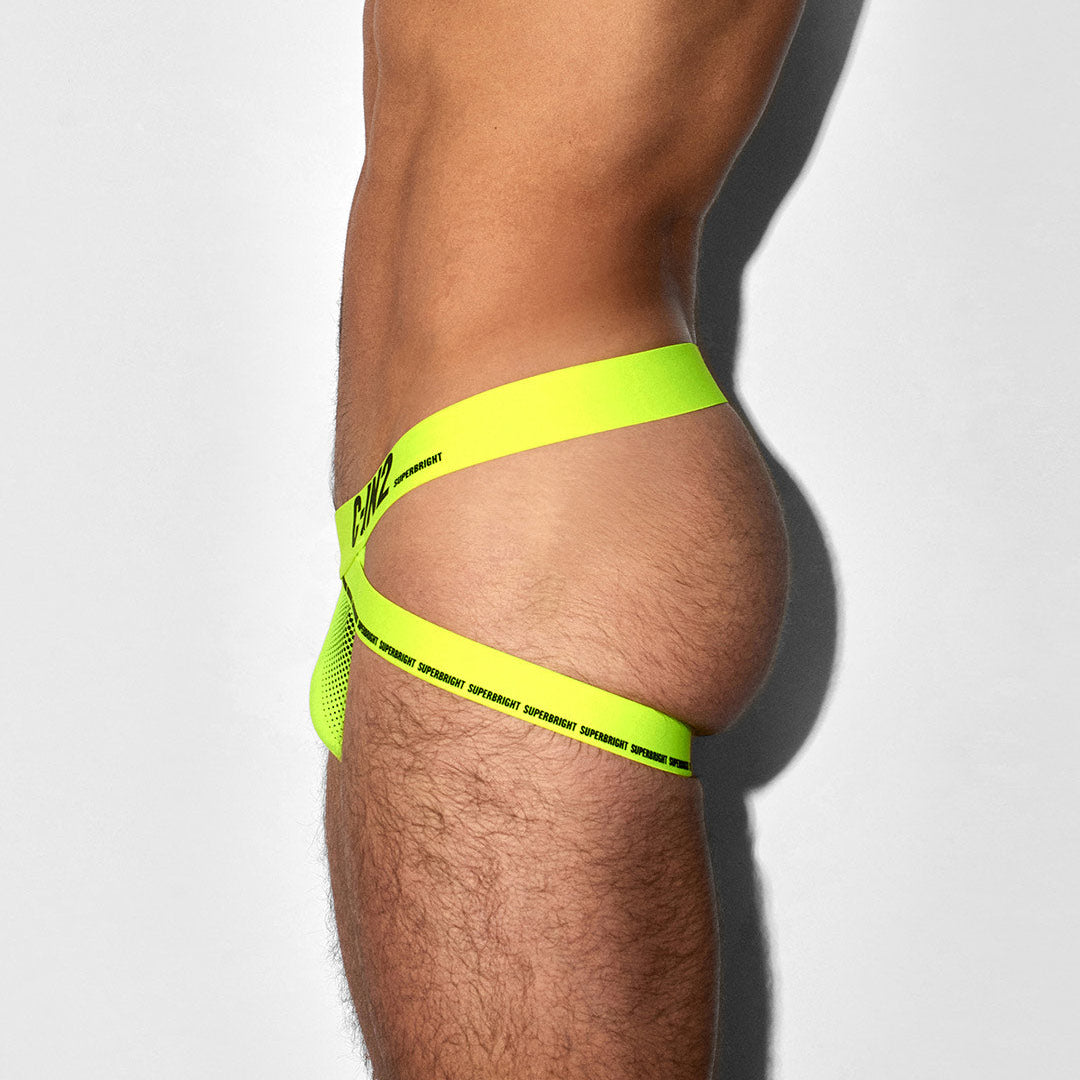 A close-up side view of a model's hip and upper thigh wearing a neon yellow-green strappy underwear or bikini-style bottom with black printed text, against a light gray studio background.