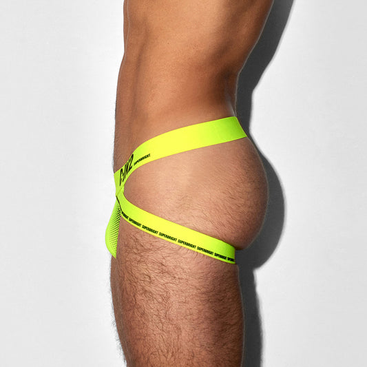 A close-up side view of a model's hip and upper thigh wearing a neon yellow-green strappy underwear or bikini-style bottom with black printed text, against a light gray studio background.