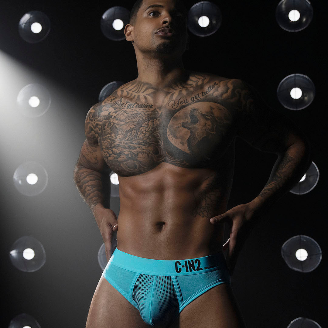 A tattooed male model wearing bright turquoise mesh briefs with black 'C-IN2' branding on the waistband, posed against a dark background with circular light fixtures.