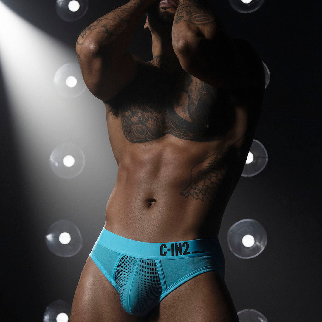 A muscular man wearing bright turquoise blue mesh briefs with the brand name 'C-IN2' printed on the front waistband, posed against a dark background with dramatic spotlights.
