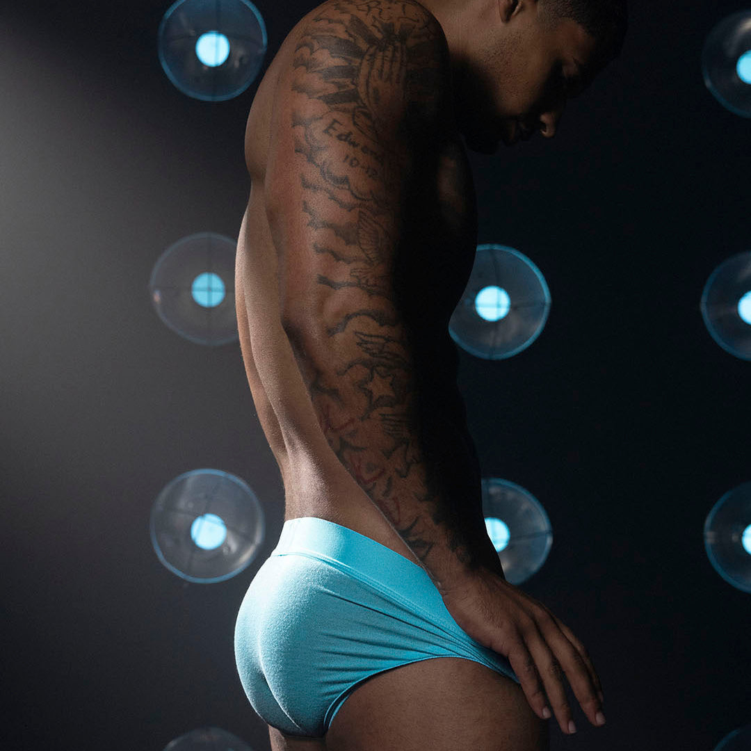 A man in side profile wearing bright turquoise blue briefs, photographed against a dark background with circular blue lights.