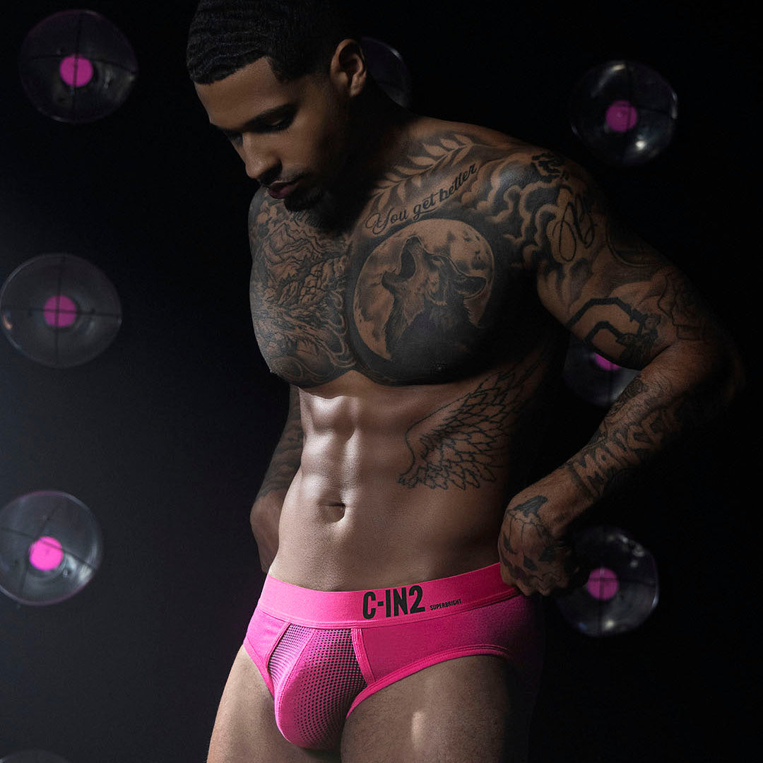 A tattooed male model wearing bright pink mesh briefs with a black waistband and the brand name 'C-IN2' printed in black, posed against a dark background with floating vinyl record graphics.
