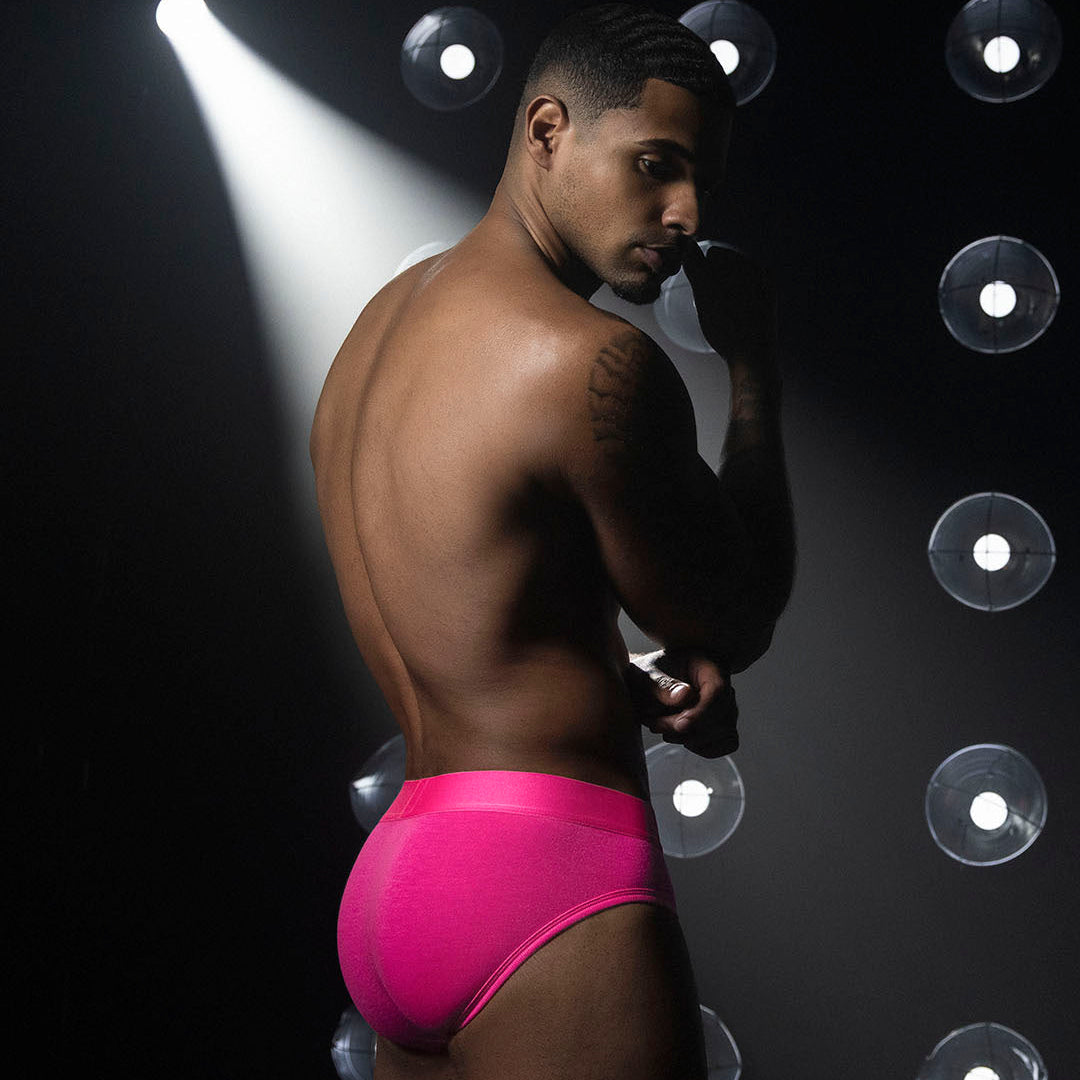 A muscular man stands in profile wearing bright hot pink briefs against a dark studio background with spotlights and circular light fixtures.