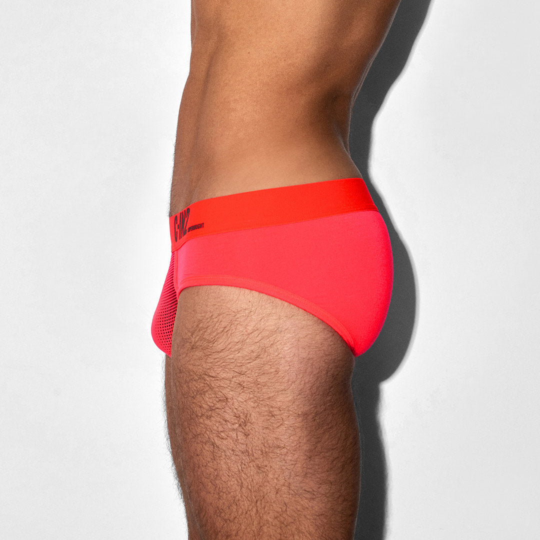 A close-up side view of a person wearing bright neon pink and orange athletic briefs against a plain light gray background.