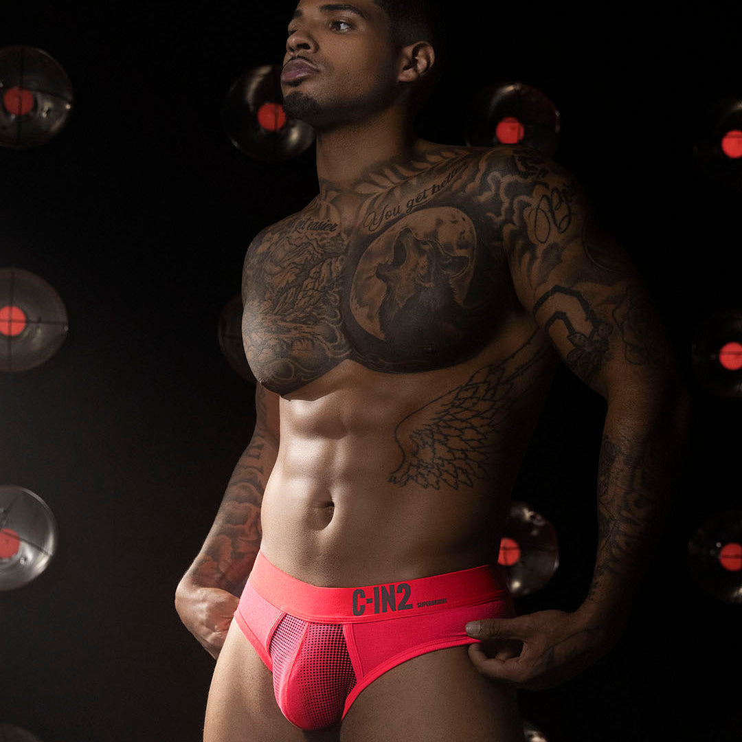 A muscular, tattooed man wearing bright pink C-IN2 men's briefs, posed against a dark background with circular light fixtures.
