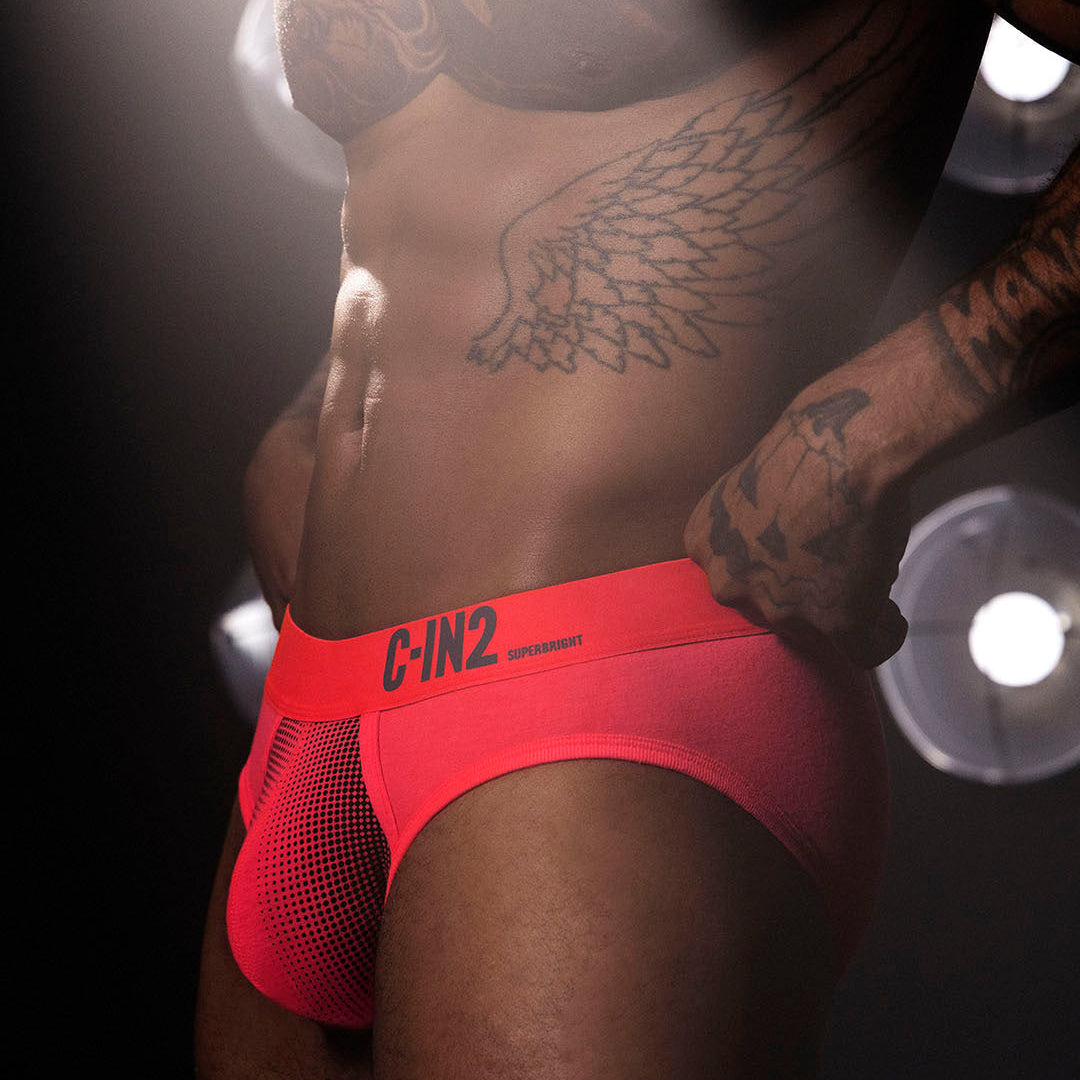A close-up lifestyle shot of a tattooed male model wearing bright neon red C-IN2 athletic briefs against a dark background with dramatic spotlighting.