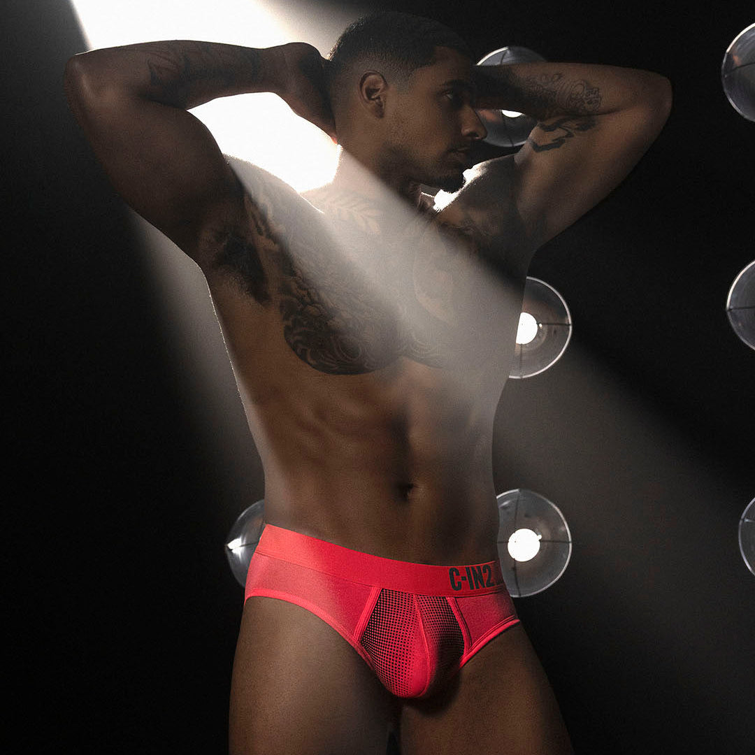 A muscular man posing in bright neon red mesh briefs against a dark background with dramatic spotlighting.